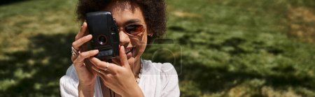 joyful african american woman taking photo on vintage camera in park in summer, banner