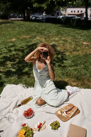 mujer afroamericana tomando fotos en cámara vintage cerca de vino y fresas, picnic de verano