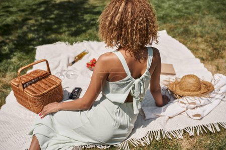 summer picnic in park, young african american woman sitting on blanket near straw basket