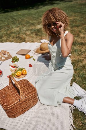 pique-nique d'été, heureuse femme afro-américaine assise près du panier de fruits, légumes et paille