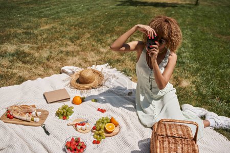 mujer afroamericana tomando fotos en cámara vintage cerca de frutas y verduras, picnic de verano