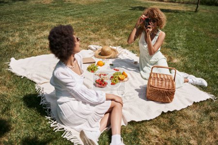 mujer afroamericana tomando fotos en cámara digital durante el picnic con su novia
