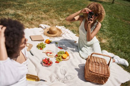 young african american woman taking photo of girlfriend on vintage camera, picnic in summer park