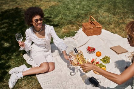 Unbekümmerte afrikanisch-amerikanische Freundinnen gießen beim Picknick Wein in die Nähe von Obst und Gemüse