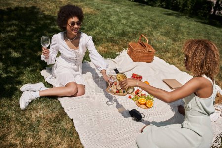 mujer afroamericana feliz sosteniendo vasos cerca de novia vertiendo vino durante el picnic de verano