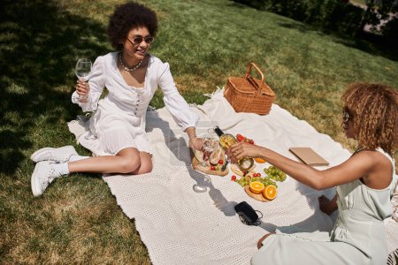 Foto de Mujer afroamericana sosteniendo copas de vino cerca de su novia, verduras y frutas, picnic de verano - Imagen libre de derechos