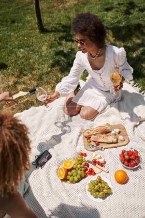 Unbekümmerte Afroamerikanerin hält Glas, während Freundin beim Sommerpicknick Wein einschenkt