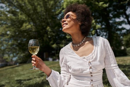 Téléchargez les photos : Femme afro-américaine insouciante et élégante dans des lunettes de soleil tenant un verre de vin dans le parc d'été - en image libre de droit