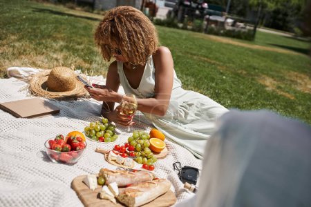 mujer afroamericana con copa de vino usando teléfono inteligente cerca de frutas y novia en el picnic en el parque