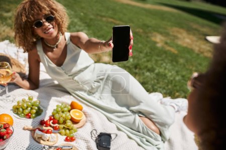 femme afro-américaine souriante tenant smartphone avec écran blanc pendant le pique-nique dans le parc d'été