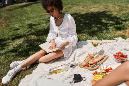 mujer afroamericana leyendo libro cerca de vino y comida en manta en parque, picnic de verano