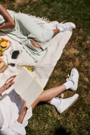 african american woman with notebook near girlfriend, fruits and wine bottle on blanket in park