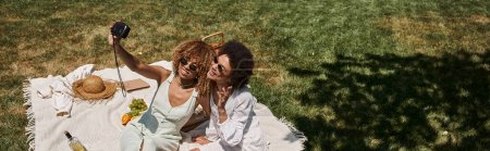 Photo for Cheerful african american girlfriends taking selfie on vintage camera on blanket near fruits in park - Royalty Free Image
