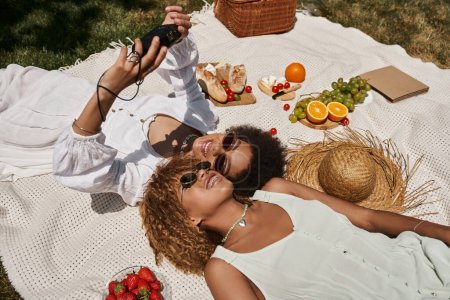afrikanisch-amerikanische Frauen machen Selfie auf Vintage-Kamera in der Nähe von Essen auf Decke, Sommerpicknick, Freude