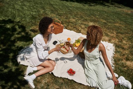 Foto de Despreocupado afroamericano novias tintineo copas de vino cerca de bocadillos en manta, picnic de verano - Imagen libre de derechos
