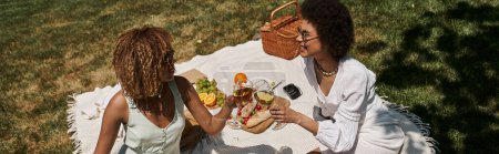 Foto de Alegres novias afroamericanas tintineando copas de vino cerca de la comida, picnic de verano en el parque, pancarta - Imagen libre de derechos