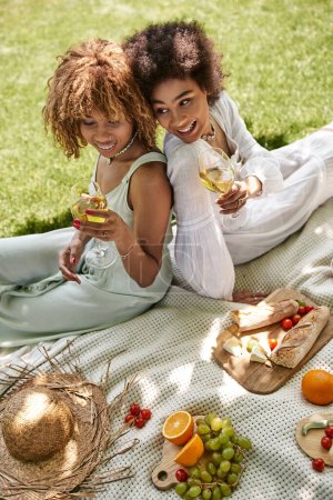 Foto de Alegres mujeres afroamericanas sentadas espalda con espalda con copas de vino cerca de comida y sombrero de paja - Imagen libre de derechos