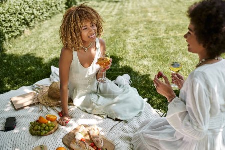 Foto de Mujer afroamericana con copa de vino sonriendo cerca de novia y comida durante el picnic de verano - Imagen libre de derechos