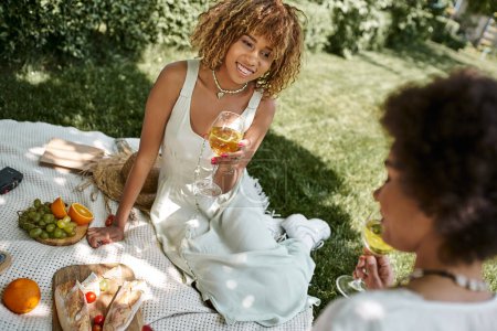 Téléchargez les photos : Joyeuse femme afro-américaine assise avec verre de vin près de la petite amie et de la nourriture pendant le pique-nique d'été - en image libre de droit