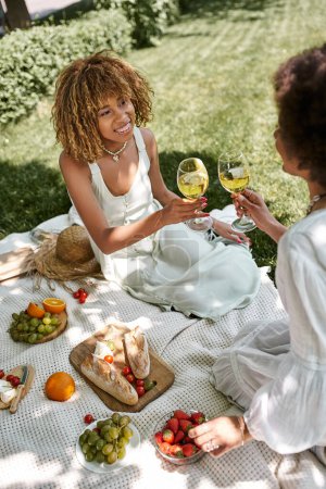Afrikanisch-amerikanische Freundinnen stoßen beim Picknick mit Weingläsern in der Nähe von Obst und Gemüse an