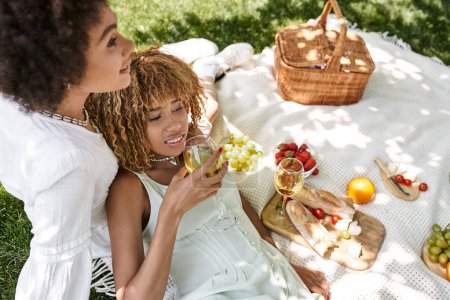 Foto de Mujer afroamericana bebiendo vino cerca de novia y bocadillos en manta, picnic de verano - Imagen libre de derechos