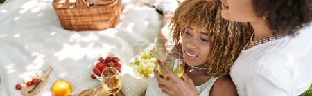 Foto de Mujer afroamericana feliz con copa de vino cerca de novia y comida en manta, ocio de verano - Imagen libre de derechos