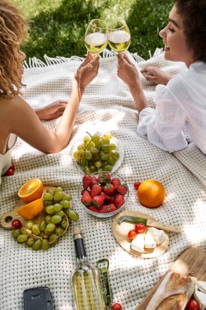 Foto de Mujeres afroamericanas tintineo copas de vino, picnic en el parque, frutas y verduras frescas en manta - Imagen libre de derechos