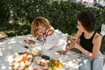 Foto de Mujer afroamericana con copa de vino hablando con su novia cerca de frutas y verduras en el picnic - Imagen libre de derechos
