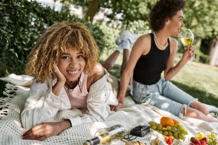 summer picnic, joyful african american woman looking at camera near girlfriend, wine and fruits