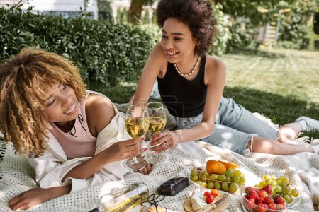 Foto de Alegres novias afroamericanas tintineando copas de vino cerca de frutas frescas, picnic de verano en el parque - Imagen libre de derechos