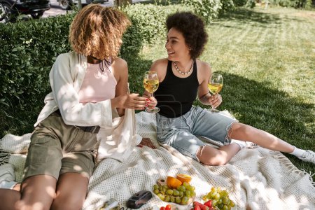 Foto de Alegre mujer afroamericana con copas de vino cerca de frutas y novia, picnic en el parque - Imagen libre de derechos
