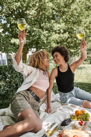 excitada mujer afroamericana brindis con copa de vino cerca de novia, disfrute, picnic en el parque