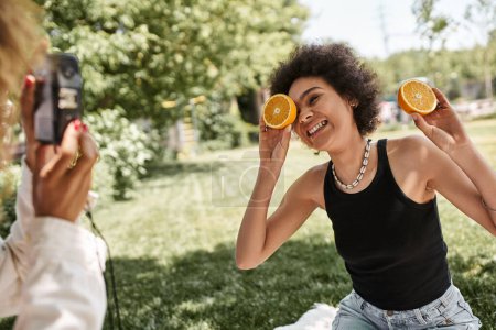 Photo for Happy african american woman posing with half orange near girlfriend with vintage camera in park - Royalty Free Image