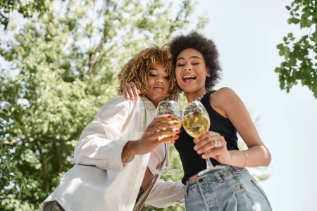 Foto de Riéndose novias afroamericanas abrazando y tintineando copas de vino, parque, verano, picnic - Imagen libre de derechos