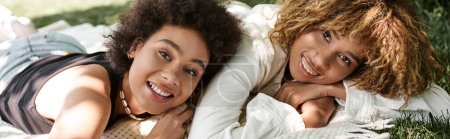 joyful african american girlfriends laying and looking at camera in park, summer happiness, banner