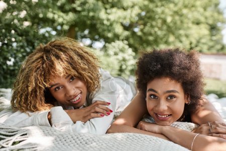 jóvenes afroamericanas novias acostadas en la manta en el parque y sonriendo a la cámara, ocio de verano