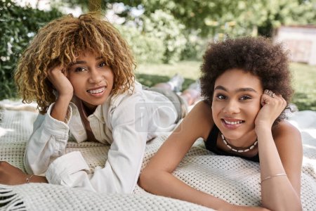 Freizeit im Sommerpark, fröhliche afrikanisch-amerikanische Freundinnen legen sich auf die Decke und schauen in die Kamera