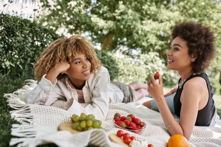 Foto de Amigas afroamericanas felices hablando cerca de frutas frescas en manta, picnic de verano en el parque - Imagen libre de derechos
