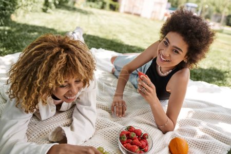 carefree african american girlfriends laying near fresh fruits on blanket, summer picnic, fun