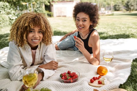novias afroamericanas jóvenes sonriendo a la cámara cerca de la comida y copas de vino, picnic de verano, parque