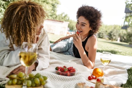 Foto de Despreocupada mujer afroamericana sosteniendo fresa y hablando con su novia en el picnic de verano - Imagen libre de derechos