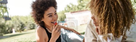sonriente mujer afroamericana con fresa hablando con su novia en el parque, picnic, pancarta