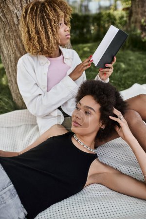 leisure in summer park, african american woman reading book near girlfriend laying on blanket