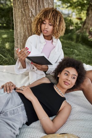 Téléchargez les photos : Africaine américaine femme lecture livre tandis que petite amie pose et regarde loin dans le parc d'été - en image libre de droit