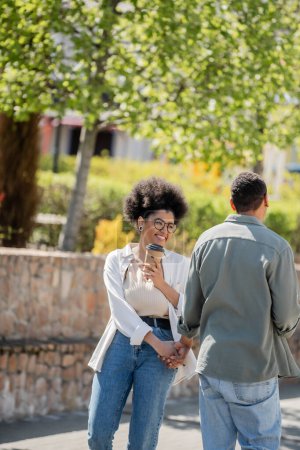 cheerful african american woman holding coffee to go and hand of boyfriend on urban street