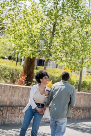 Téléchargez les photos : Positive femme afro-américaine tenant du café pour aller près de petit ami dans la rue urbaine - en image libre de droit