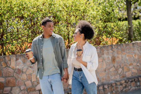sonriente pareja afroamericana sosteniendo café para ir mientras habla y camina por la calle urbana