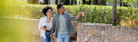 Téléchargez les photos : Souriant homme afro-américain avec du café pour aller pointant vers la rue urbaine près de petite amie, bannière - en image libre de droit