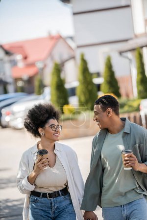 sonriente pareja afroamericana con café para llevar cogido de la mano en la calle urbana