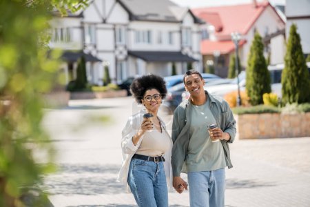 Téléchargez les photos : Femme afro-américaine souriante avec café pour aller marcher avec petit ami près des maisons dans la rue urbaine - en image libre de droit